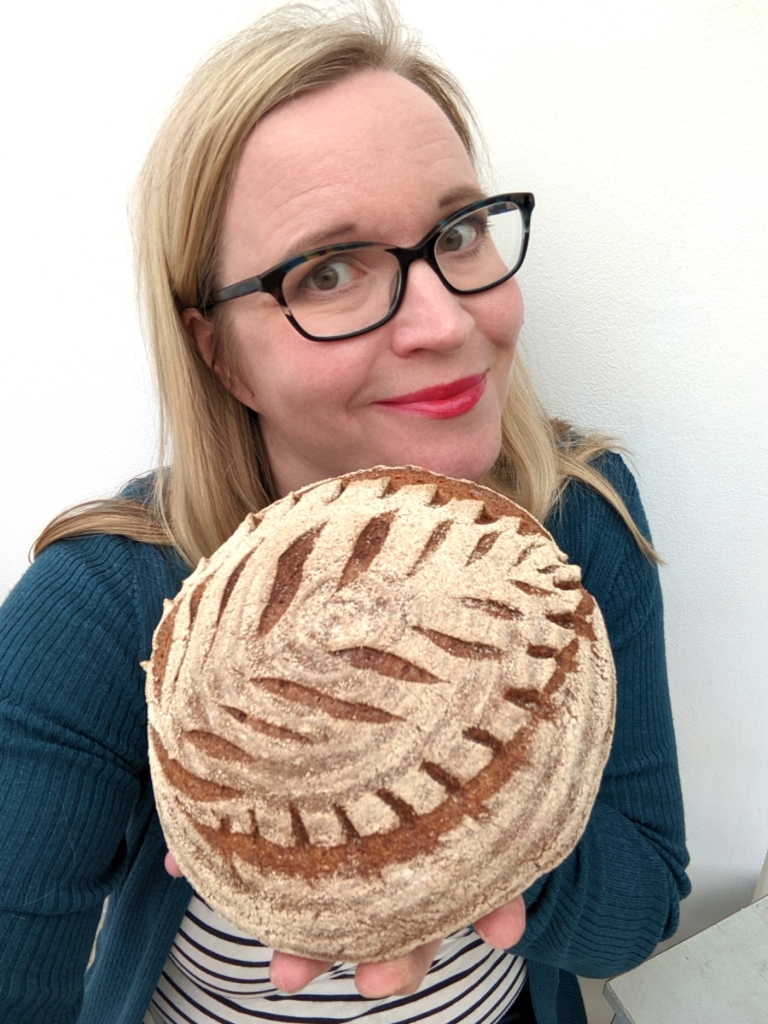 A woman with glasses, red lipstick and blonde long hair wearing a teal cardigan and stripey t-shirt holding a round loaf of bread with a leaf pattern scored into it.