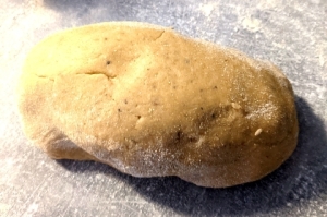 A lump of gluten-free cardamom bun dough on a grey floured worktop. Specs of black cardamom visible in the dough.