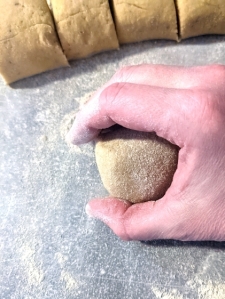 Piece of gluten-free cardamom bun dough in a cupped kaukasian hand forming a ball against the gray floured worktop with pieces of dough placed in a row in the background.