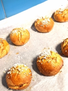 Smaller gluten-free baked golden brown cardamom buns on a baking tray lined with non-stick baking paper with a turquioise Metro tile at the background. The buns have clear cracks and pearl sugar on top.
