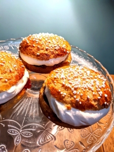 Gluten-free Finnish Shrove buns topped with pearl sugar and filled with whipped cream and strawberry jam in between the halved buns. The buns sit on a glass platter on a wooden table with a duck egg coloured wall at the background.