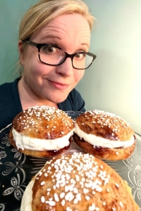 Kaukasian blonde lady with tortoise pattern glasses and teal cardigan holding a glass platter of large gluten-free cardamom buns topped with pearl sugar and filled with whipped cream and strawberry jam placed in between the two halves of the bun.