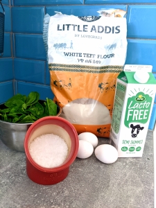 The ingredients of the gluten-free spinach pancakes on a mock concrete kitchen worktop with a turquioise metro tile splashback in the backgroud. Little Addis by Lovegrass white teff flour in a large plastic bag, Arla Lacto Gree milk carton, baby spinach in a steel bowl, three white chicken eggs and a terracotta coloured salt container with Maldon finger salt in the foreground.