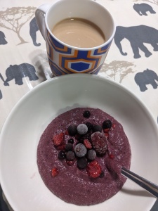 A mug of milky coffee and a white bowl with purple whipped berry porridge in topped with frozen berries.