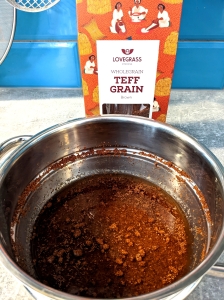 A packet of brown Lovegrass wholegrain teff grain  and a saucepan with teff grain soaking in water on a countertop with utensils and a teal tile wall behind.