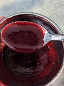 Smooth dark red berry puree which has been passed through a sieve in a steal spoon - a steel bowl with puree is seen underneath