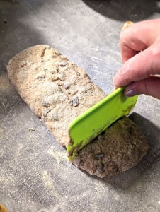 An oblong of gluten-free hot cross bun dough on a floured workthop being cut to pieces using a green dough scraper