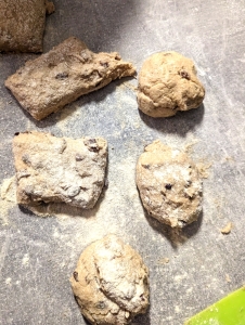 Six pieces of gluten-free hot cross bun mixture, three of which have been formed to round buns on a floured worktop.