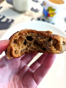 A hand holding half a hot cross bun with the inside showing large holes, candied peel and raisins.