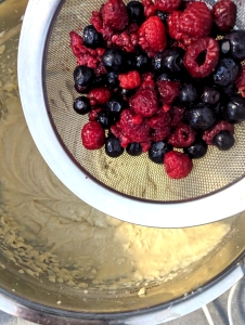 Drained defrosted raspberries and blueberries in a steel sieve poised on top of a steel bowl with the fluffy gluten-free cake batter underneat ready to go in.
